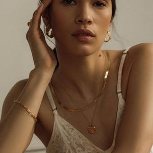 May include: A gold jewelry set consisting of a flat herringbone chain necklace, a delicate pendant necklace with a textured circular charm, a thin link bracelet, and a pair of chunky, irregular hoop earrings with a hammered, organic finish.