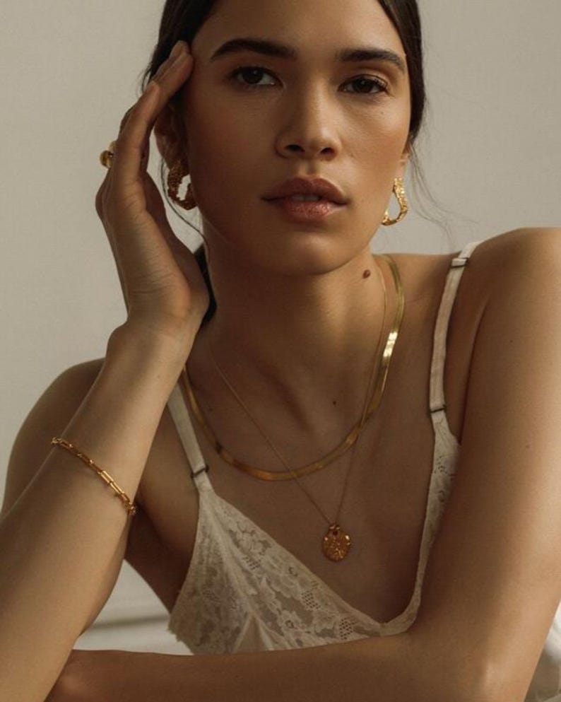 May include: A gold jewelry set consisting of a flat herringbone chain necklace, a delicate pendant necklace with a textured circular charm, a thin link bracelet, and a pair of chunky, irregular hoop earrings with a hammered, organic finish.