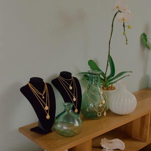 May include: Two black velvet jewelry display busts featuring layered gold-tone necklaces with circular pendants. Beside them are two green glass vases, one white ribbed vase, and a potted orchid, all arranged on a wooden shelf.