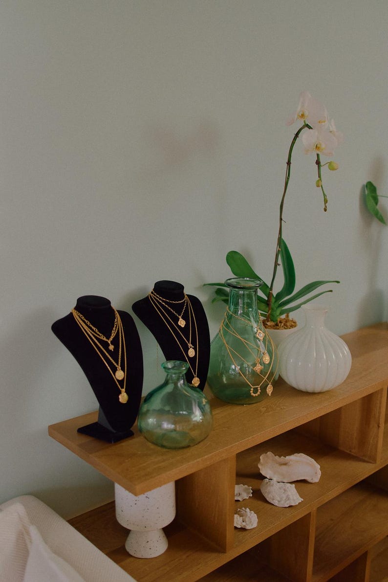 May include: Two black velvet jewelry display busts featuring layered gold-tone necklaces with circular pendants. Beside them are two green glass vases, one white ribbed vase, and a potted orchid, all arranged on a wooden shelf.