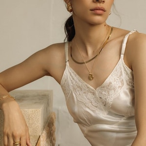 May include: A white satin camisole featuring delicate lace trim along the V-neckline, paired with gold jewelry including a layered necklace with a round pendant, textured gold stud earrings, a thin chain bracelet, and a stacked gold ring.