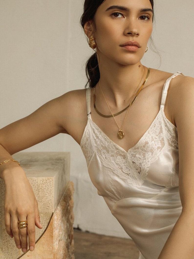 May include: A white satin camisole featuring delicate lace trim along the V-neckline, paired with gold jewelry including a layered necklace with a round pendant, textured gold stud earrings, a thin chain bracelet, and a stacked gold ring.