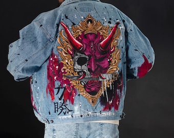 May include: Light blue denim jacket with a vibrant, hand-painted design on the back. The artwork features a detailed, stylized oni mask in shades of red, purple, and gold, with Japanese characters. Splatter paint adds a unique touch.