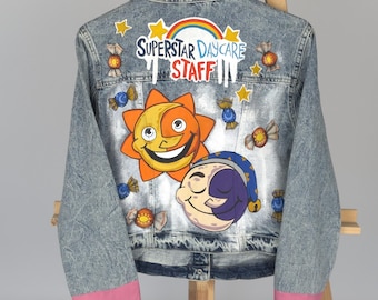 May include: A denim jacket with a colorful design featuring a smiling sun and moon, candy, and the text "SUPERSTAR DAYCARE STAFF". The jacket has pink trim on the sleeves and is displayed on a wooden easel.
