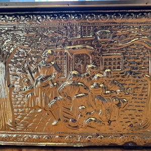Può includere: Un forziere rettangolare in ottone con un rilievo in rilievo raffigurante un raduno di figure in un ambiente di villaggio. L'opera d'arte dettagliata comprende alberi, edifici e persone, il tutto reso in una calda tonalità dorata.