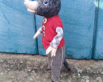 May include: A handmade felt mole figurine wearing a red vest with wooden buttons, plaid sleeves, and tweed pants. The mole has a black head, a pink nose, and is standing on a concrete surface in front of a blue door.