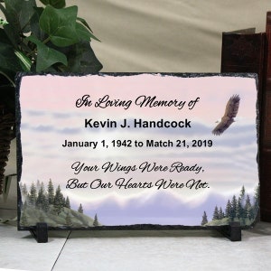 May include: A black slate memorial plaque with a mountain and eagle image. The text reads "In Loving Memory of Kevin J. Handcock January 1, 1942 to March 21, 2019 Your Wings Were Ready, But Our Hearts Were Not."
