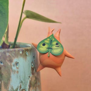 以下が含まれることがあります： ゼルダの伝説シリーズのキャラクター、コログの木製フィギュア。オレンジ色のフィギュアは緑の葉のマスクをしており、青緑色の陶器の鉢の縁にちょこんと座っています。鉢はまだら模様です。