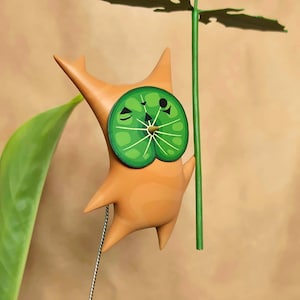 May include: A whimsical wooden sculpture of a creature with a green lily pad face and a green leaf umbrella. The sculpture is primarily tan with green accents and a silver pull string. The design is reminiscent of a cartoon character.