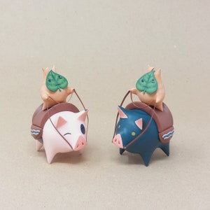 以下が含まれることがあります： 葉の形をした帽子をかぶった小さなセラミック製の豚の置物2つ。左側の豚はピンクで、右側の豚は青です。両方の豚は茶色の鞍をつけており、手綱を持っています。