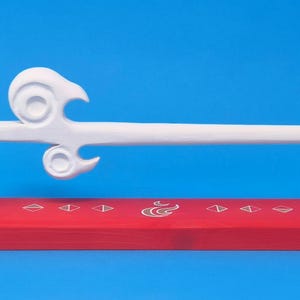 May include: A white sword replica with a stylized, curved hilt design, mounted horizontally on two green supports. The supports rest on a rectangular red wooden base featuring white geometric symbols and a central flame-like icon.