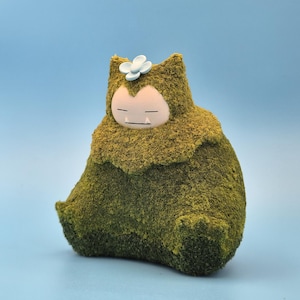 Op de afbeelding: Een met mos bedekt Snorlax-beeldje met een lichtblauwe bloem op zijn hoofd. Het beeldje heeft een lichtroze gezicht met gesloten ogen en een kleine mond. De achtergrond is effen lichtblauw.