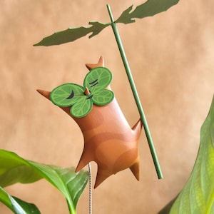 May include: A whimsical brown and green figurine of a creature with a leafy head and a long green stem. The creature is holding onto the stem with its arms and has a happy expression on its face.
