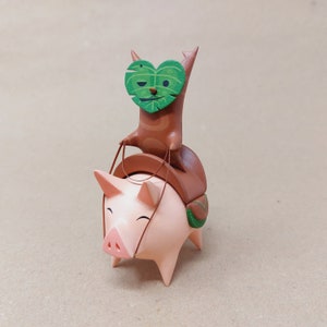 以下が含まれることがあります： 手描きの木製の豚の置物で、茶色と緑の猫が背中に乗っています。猫は顔に緑の葉の形をしたマスクをしています。