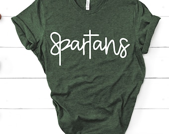 May include: A dark green t-shirt with the word "Spartans" in white script across the front. The shirt is made of a soft material and has rolled-up sleeves. The shirt is displayed on a white wooden surface.