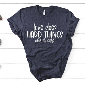 May include: Navy blue t-shirt with the text "love does HARD THINGS" and the hashtag "#fostercare" in white. The shirt is made of a soft material and has rolled-up sleeves. The shirt is displayed on a white wooden surface.