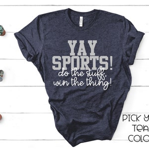 May include: Navy blue t-shirt with the text "YAY SPORTS! do the stuff, win the thing!" in gray and white. The shirt also has the text "PICK YOUR TEAM COLORS!" in black. The shirt is displayed on a white wooden surface.