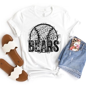 May include: White t-shirt with a black and white baseball design and the word "BEARS". The baseball graphic has a leopard print pattern. The shirt is paired with denim shorts, a floral phone case, and white sandals.