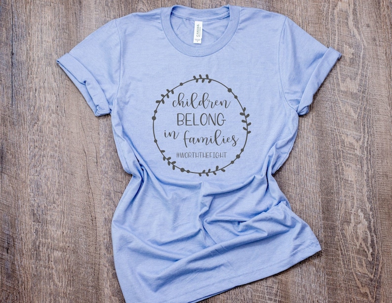 May include: Light blue t-shirt with rolled-up sleeves. The shirt features a circular design with the words "children belong in families" and the hashtag "#WORTHYTHEFIGHT" in a dark gray color.