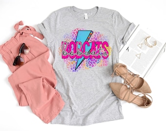 May include: A heather gray t-shirt with the word "BOBCATS" in a pink and black font, with a blue lightning bolt design. The shirt is surrounded by a colorful leopard print design. The image also includes pink pants, sunglasses, and tan flats.