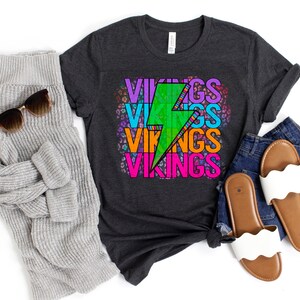 May include: Dark gray t-shirt with the word "VIKINGS" stacked in colorful text with a green lightning bolt graphic. The shirt is styled with a gray sweater, sunglasses, blue jeans, and white sandals.