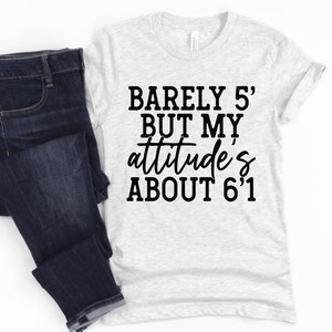 May include: A light gray t-shirt with the text "Barely 5' but my attitude's about 6'1" in black script. The shirt is paired with dark blue denim jeans. The t-shirt is a casual wear item.
