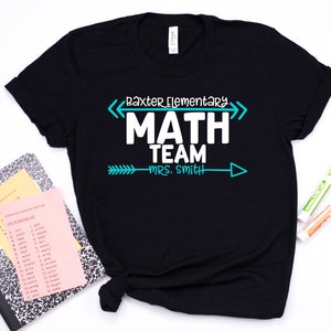May include: Black t-shirt with white text that reads "Baxter Elementary MATH TEAM Mrs. Smith" in a teal arrow design. The shirt is next to a notebook, papers, and markers.