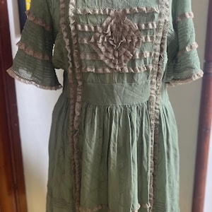 Könnte beinhalten: Ein salbeigrünes Kleid mit kurzen Ärmeln und rundem Ausschnitt. Das Kleid hat eine aufwendige, hellbraune Spitzenborte am Oberteil, an den Ärmeln und am Rock. Eine dekorative florale Verzierung befindet sich in der Mitte des Oberteils.