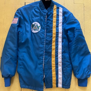 Könnte beinhalten: Eine blaue Vintage-Jacke mit schwarzem Kragen und einem weißen und gelben Streifen auf der Vorderseite. Ein amerikanischer Flaggenaufnäher befindet sich auf dem linken Ärmel und ein runder Aufnäher auf der Brust. Die Jacke hat einen Reißverschluss und blaue Bündchen.