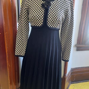 May include: A vintage black and white checkered jacket and dress set. The jacket has long sleeves and a small flower detail. The dress is black with a pleated skirt. The outfit is displayed on a mannequin.