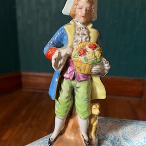 May include: A porcelain figurine of a man in 18th-century attire, holding a basket of roses. The figure wears a blue coat with yellow trim, green pants, and a white hat. A small dog stands at his feet.