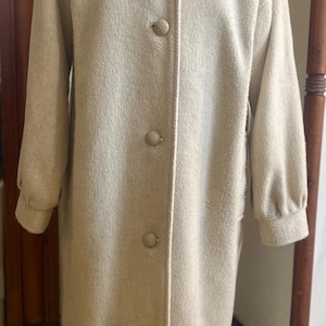 May include: A long, light beige coat with a high collar and large buttons. The coat has long sleeves and a tailored fit, suitable for cooler weather. The coat is made of a soft, textured fabric.