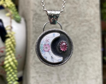 Può includere: Una collana con catena d'argento con un ciondolo a forma di mezzaluna. La luna è in porcellana bianca con un motivo floreale e una pietra preziosa rosa al centro.