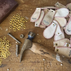 Peut inclure: Gros plan sur une vari&eacute;t&eacute; de patchs en cuir avec des coutures rouges et le mot "Rawlings" imprim&eacute; dessus. Les patchs sont dispos&eacute;s sur une surface en bois avec un marteau, des &oelig;illets et un morceau de tissu brun.