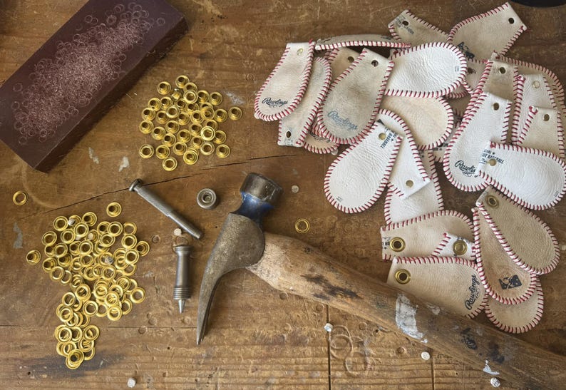 Peut inclure: Gros plan sur une vari&eacute;t&eacute; de patchs en cuir avec des coutures rouges et le mot "Rawlings" imprim&eacute; dessus. Les patchs sont dispos&eacute;s sur une surface en bois avec un marteau, des &oelig;illets et un morceau de tissu brun.