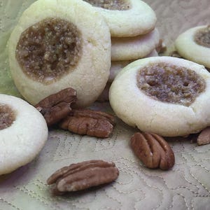 May include: Close-up of several round cookies with a light-colored exterior and a brown, nut-filled center. Several pecan halves are scattered around the cookies. The cookies are arranged on a light-colored, textured surface.