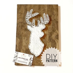 Könnte beinhalten: Weiße String Art eines Hirschkopfes auf braunem Holzgrund. Die Kunst ist gerahmt und hat eine Visitenkarte mit dem Text "Kiki String Art by Chiara Mai" und einer Website-Adresse. Der Text "DIY PATTERN" ist ebenfalls auf dem Bild zu sehen.