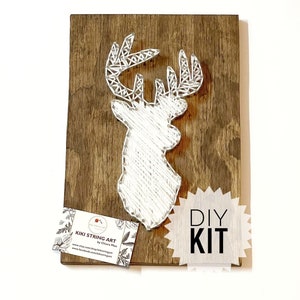 May include: DIY string art kit featuring a white deer head design on a brown wooden base. The kit includes all the materials needed to create the art, including string, nails, and a wooden base. The kit is perfect for beginners and experienced crafters alike.