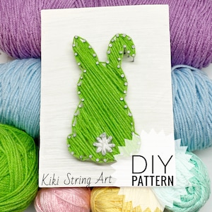 Op de afbeelding: DIY string art patroon met een groene konijn met een witte bloem. Het patroon is omgeven door bollen garen in verschillende kleuren.