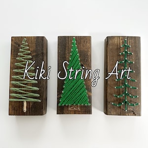 May include: Three wooden signs with string art of green Christmas trees. The signs are arranged horizontally and have the text "Kiki String Art" in the center.