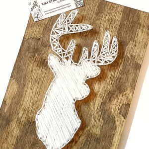May include: White string art of a deer head on a brown wooden background. The art is titled "Kiki String Art" by Chiara Mau.
