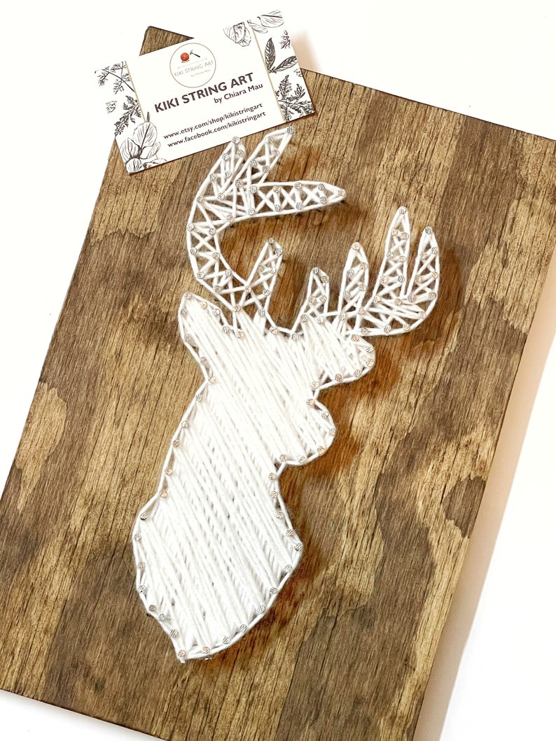 May include: White string art of a deer head on a brown wooden background. The art is titled "Kiki String Art" by Chiara Mau.