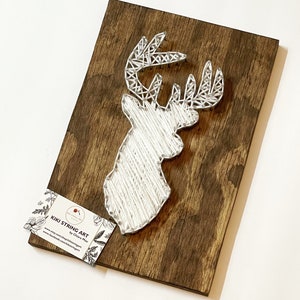 May include: White string art of a deer head on a brown wooden background. The art is titled "Kiki String Art" by Chiara Maru. The website address is www.etsy.com/shop/kikistringart and the Facebook address is www.facebook.com/kikistringart.