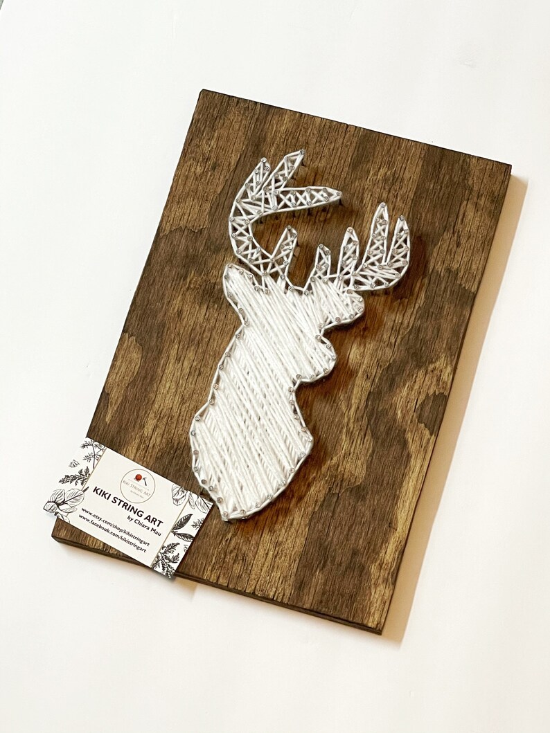 May include: White string art of a deer head on a brown wooden background. The art is titled "Kiki String Art" by Chiara Maru. The website address is www.etsy.com/shop/kikistringart and the Facebook address is www.facebook.com/kikistringart.