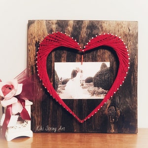 May include: Red string art heart with a wedding photo inside, on a dark wood background. The text "Kiki String Art" is at the bottom of the frame.