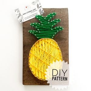 Op de afbeelding: DIY string art patroon van een ananas met geel en groen garen op een houten plank. De tekst "DIY PATTERN" staat op de plank.