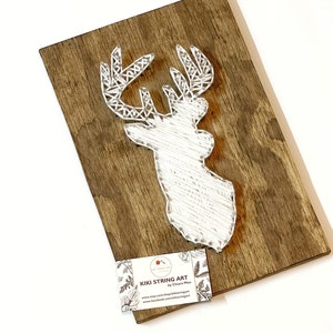 May include: White string art of a deer head on a wooden background. The art is signed "Kiki String Art by Chiara Mai" with a website and Facebook page listed.
