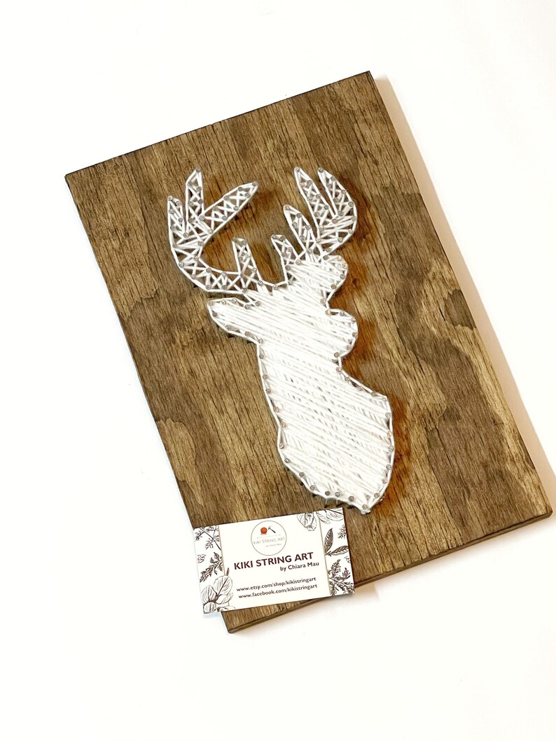 May include: White string art of a deer head on a wooden background. The art is signed "Kiki String Art by Chiara Mai" with a website and Facebook page listed.