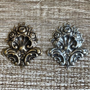 May include: Two ornate metal embellishments with a floral design. One is silver and the other is gold.