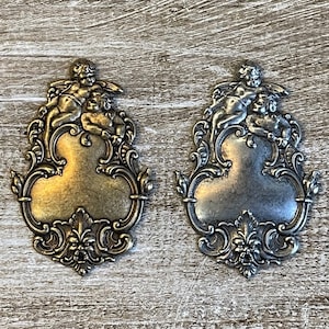 May include: Two ornate metal charms featuring cherubs in a playful embrace. The charms are in a gold and silver finish, with intricate details and a blank space in the center.